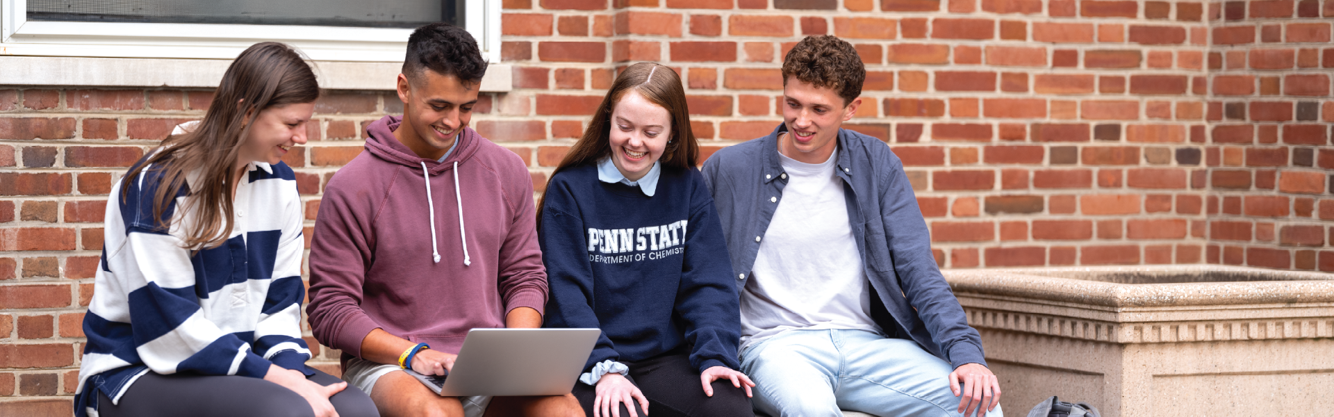Group of students sitting around laptop on exterior steps in front of brick building.