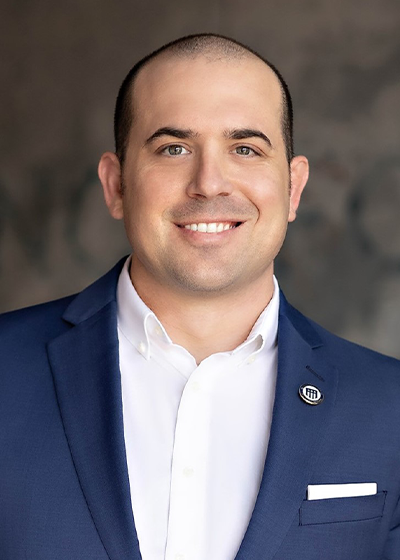 Ethan Merrill smiling in a blue suit with a white shirt, against a blurred wall background.