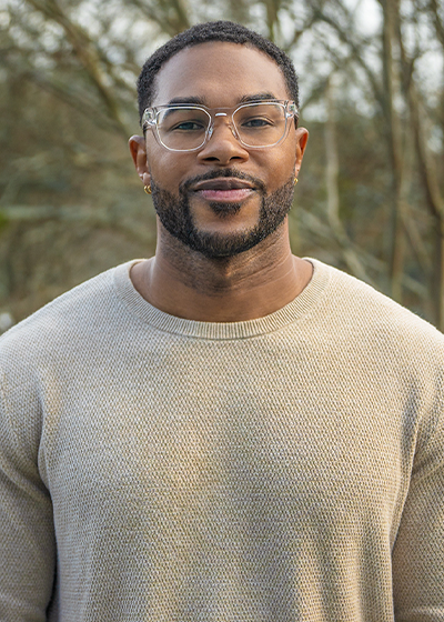 Rubain Henry wearing glasses and a beige sweater stands outdoors with trees in the background.