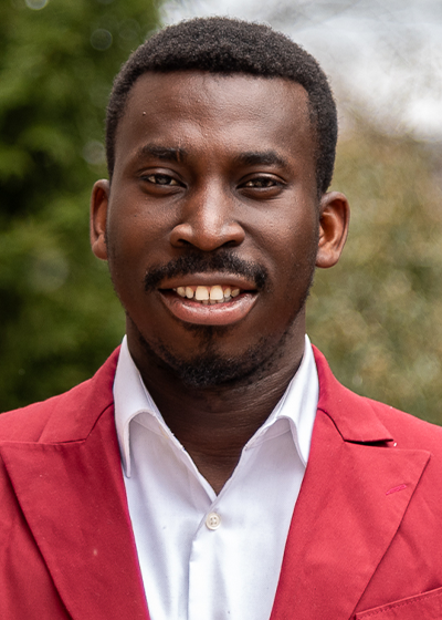 Patrick Sarpong wearing a red blazer and white shirt, standing outdoors with greenery in the background.