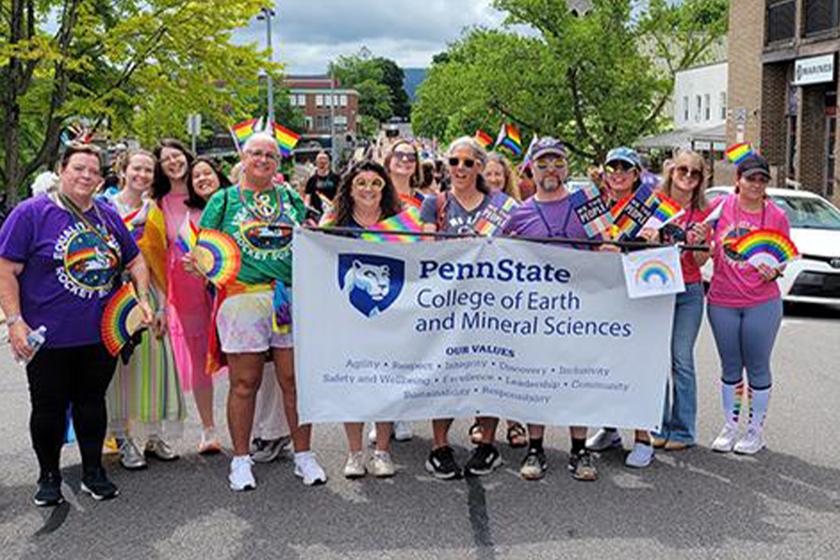 Group in multi-colored clothes standing outside holding Penn State College of Earth and Mineral Sciences banner. 