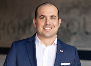 Ethan Merrill smiling in a blue suit with a white shirt, against a blurred wall background.