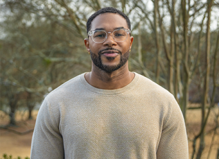 Rubain Henry wearing glasses and a beige sweater stands outdoors with trees in the background.